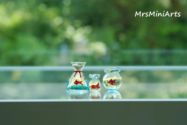 May include: Three miniature glass fish bowls, each containing a red fish. The bowls vary in shape: a bag, a small jar, and a round bowl. The bag-shaped bowl has a red ribbon. The background is a blurred green.