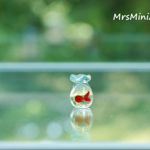 May include: A miniature glass fishbowl with a red fish inside. The bowl has a light blue ruffled top and is filled with clear liquid. The image is set against a blurred green background, with the text "MrsMiniArts" in the upper right corner.