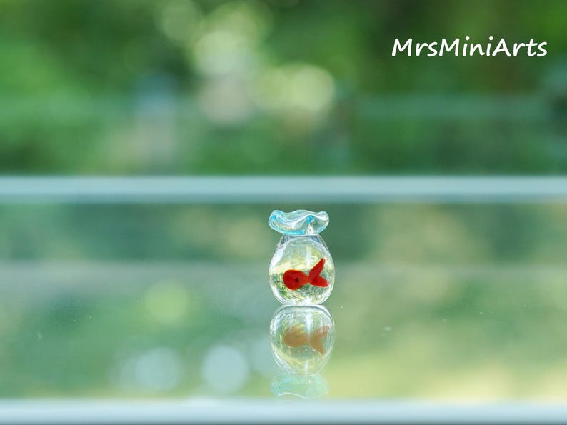 May include: A miniature glass fishbowl with a red fish inside. The bowl has a light blue ruffled top and is filled with clear liquid. The image is set against a blurred green background, with the text "MrsMiniArts" in the upper right corner.