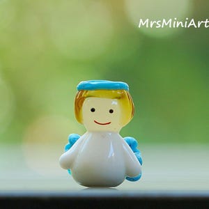May include: A small, white figurine of an angel with a blue halo and wings. The angel has yellow hair and a smiling face. The text "MrsMiniArts" is visible in the background.