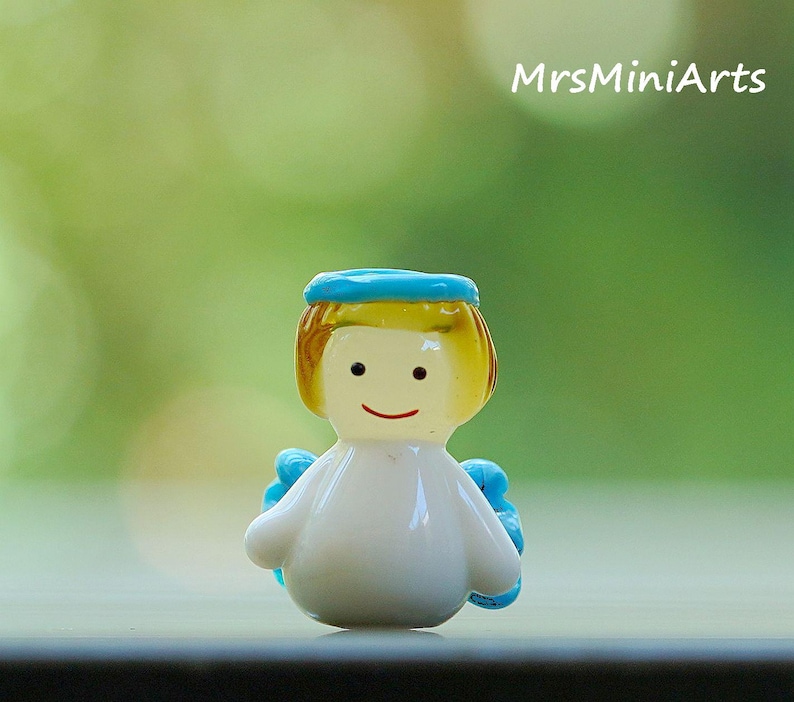 May include: A small, white figurine of an angel with a blue halo and wings. The angel has yellow hair and a smiling face. The text "MrsMiniArts" is visible in the background.