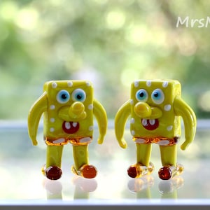 May include: Two small glass figurines of a cartoon character. The figurines are yellow with white spots and brown legs. The character has a big smile and blue eyes. The figurines are on a white surface.
