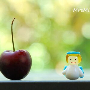 May include: A ripe, dark red cherry with a long green stem stands next to a small, smiling angel figurine. The angel has yellow hair, a blue halo, and blue wings. The background is a blurred green.