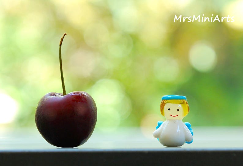May include: A ripe, dark red cherry with a long green stem stands next to a small, smiling angel figurine. The angel has yellow hair, a blue halo, and blue wings. The background is a blurred green.