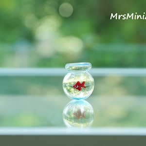 May include: A miniature glass fishbowl with a red fish inside, reflected below. The fishbowl is clear with a light blue rim. The background is a blurred green. The text "MrsMiniArts" is in the upper right corner.