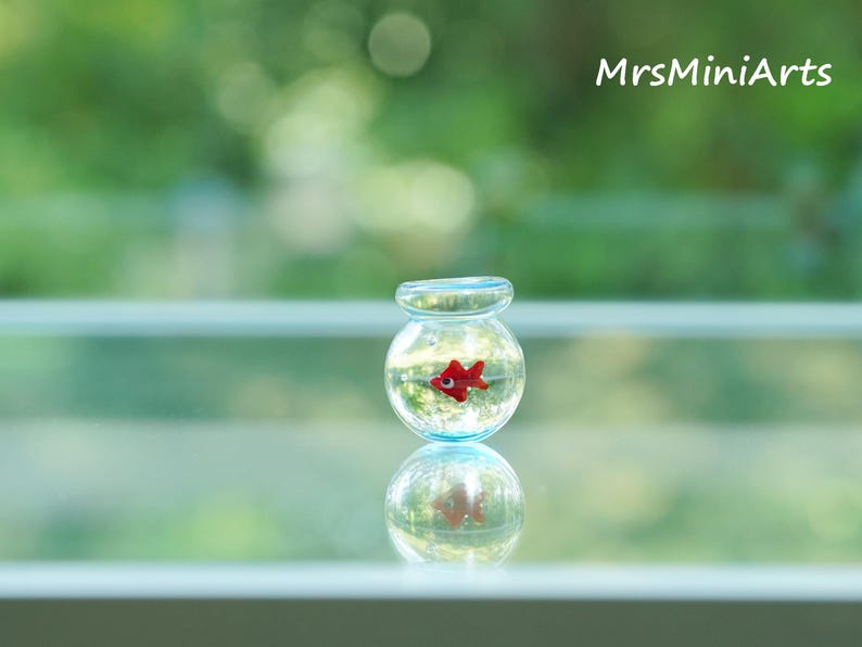 May include: A miniature glass fishbowl with a red fish inside, reflected below. The fishbowl is clear with a light blue rim. The background is a blurred green. The text "MrsMiniArts" is in the upper right corner.