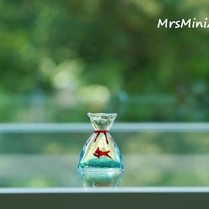 May include: A miniature glass sculpture of a fish in a bag. The clear glass bag has a red ribbon tie and a red fish inside. The sculpture is set against a blurred green background, with the text "MrsMiniArts" in the upper right corner.