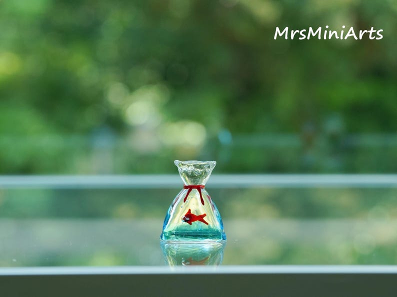May include: A miniature glass sculpture of a fish in a bag. The clear glass bag has a red ribbon tie and a red fish inside. The sculpture is set against a blurred green background, with the text "MrsMiniArts" in the upper right corner.