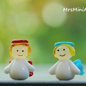 May include: Two small, glass angel figurines. One angel has a red halo and wings, the other has a blue halo and wings. Both have yellow hair, smiling faces, and white bodies. The background is a soft green.