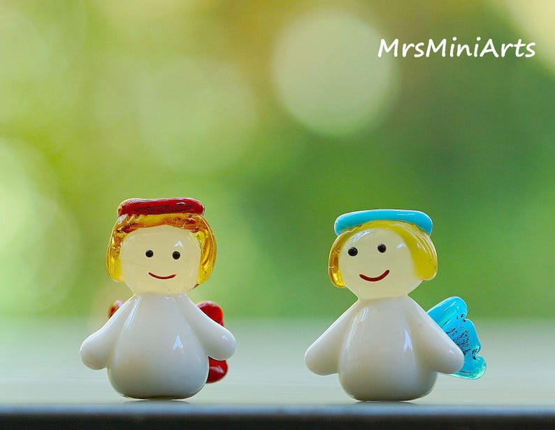 May include: Two small, glass angel figurines. One angel has a red halo and wings, the other has a blue halo and wings. Both have yellow hair, smiling faces, and white bodies. The background is a soft green.