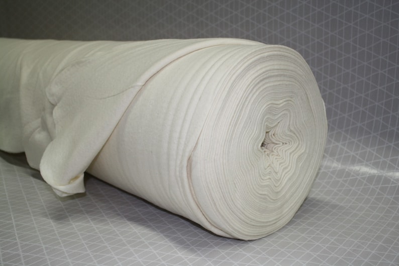 May include: A roll of off-white fabric, tightly wound, with visible layers. The fabric appears soft and possibly felted, with a slight sheen. The roll is positioned horizontally, showcasing the texture and volume of the material. The background is a light gray with a geometric pattern.