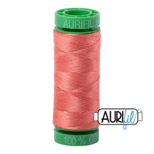 May include: A spool of coral-colored Aurifil thread. The thread is tightly wound on a spool with green caps. The top cap has the brand name "AURIFIL" printed on it. The bottom cap reads "100% CO".