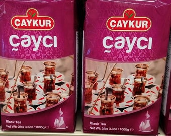 May include: A magenta-coloured bag of Caykur Cayci black tea. The packaging features the brand logo, the text Cayci, and a graphic of multiple small glass cups filled with dark tea on white saucers. The net weight is listed as 1000g.