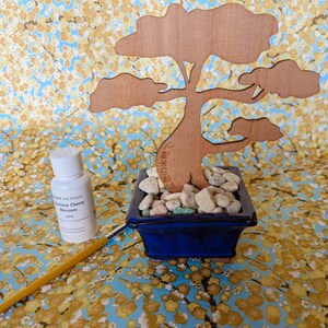 May include: A wooden bonsai tree cutout in a blue ceramic pot with white stones. A small bottle of Japanese Cherry Blossom scent is next to a yellow paintbrush.