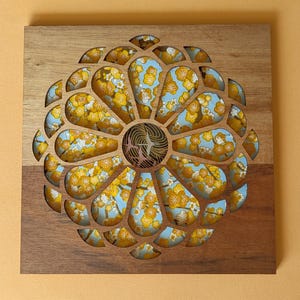May include: Wooden wall art with a floral design. The artwork features a circular pattern cut out of the wood, revealing a blue fabric with yellow floral patterns. The wood has a natural, light brown color.