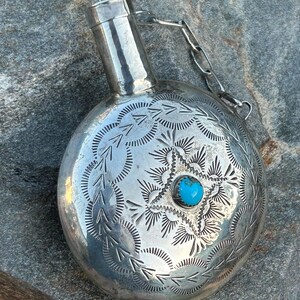 Puede incluir: Una petaca plateada con intrincados patrones grabados y una piedra turquesa en el centro. La petaca tiene un cuerpo redondo, un cuello cil&iacute;ndrico y una cadena sujeta a la tapa. El dise&ntilde;o grabado presenta motivos de estrella y hojas.