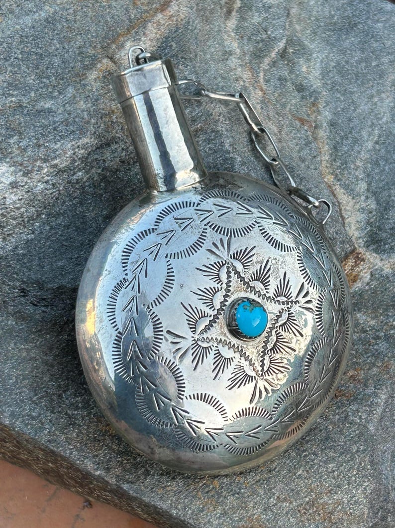 Puede incluir: Una petaca plateada con intrincados patrones grabados y una piedra turquesa en el centro. La petaca tiene un cuerpo redondo, un cuello cil&iacute;ndrico y una cadena sujeta a la tapa. El dise&ntilde;o grabado presenta motivos de estrella y hojas.