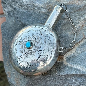 Puede incluir: Una petaca plateada con intrincados dise&ntilde;os grabados y una peque&ntilde;a piedra turquesa en el centro. La petaca tiene un cuerpo redondeado, un cuello largo y una cadena sujeta a la tapa. La petaca es probablemente de metal.
