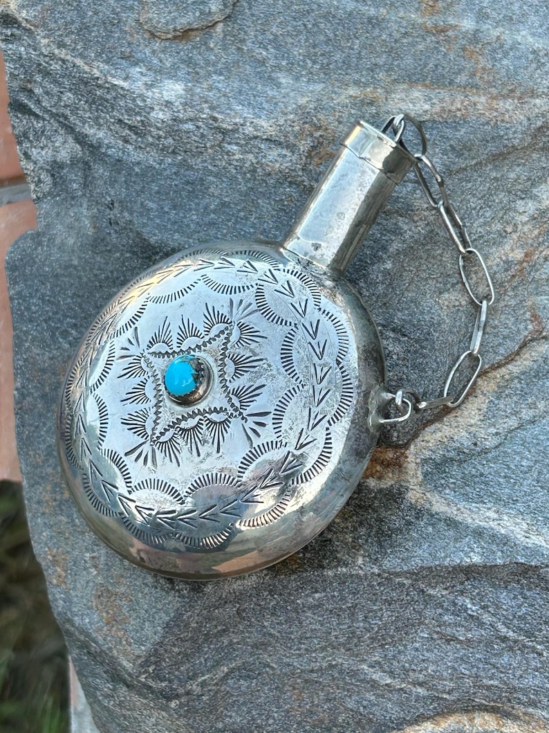 Puede incluir: Una petaca plateada con intrincados dise&ntilde;os grabados y una peque&ntilde;a piedra turquesa en el centro. La petaca tiene un cuerpo redondeado, un cuello largo y una cadena sujeta a la tapa. La petaca es probablemente de metal.