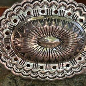 May include: Close-up of a silver-colored belt buckle with an oval center and intricate, detailed patterns. The buckle is set against a brown leather belt, showcasing a Southwestern or Western design.