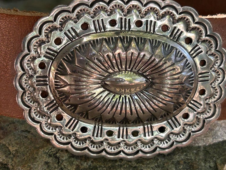 May include: Close-up of a silver-colored belt buckle with an oval center and intricate, detailed patterns. The buckle is set against a brown leather belt, showcasing a Southwestern or Western design.