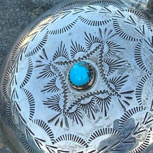 Puede incluir: Primer plano de una petaca redonda plateada con intrincados patrones grabados. Una vibrante piedra turquesa est&aacute; engastada en el centro. La petaca tiene un asa y probablemente est&aacute; hecha de metal.
