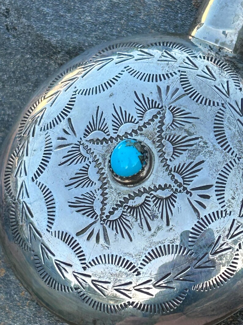 Puede incluir: Primer plano de una petaca redonda plateada con intrincados patrones grabados. Una vibrante piedra turquesa est&aacute; engastada en el centro. La petaca tiene un asa y probablemente est&aacute; hecha de metal.