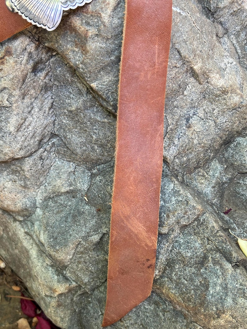 May include: Close-up of a brown leather belt with a silver buckle. The belt is made of a single strip of leather and shows natural wear. The silver buckle has a decorative design.