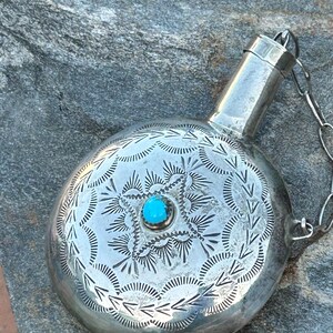 Puede incluir: Una petaca de plata con una piedra turquesa en el centro. La petaca tiene un cuerpo redondo con patrones grabados y un peque&ntilde;o pico con una cadena. La petaca es probablemente de plata de ley.