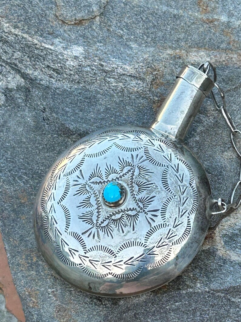 Puede incluir: Una petaca de plata con una piedra turquesa en el centro. La petaca tiene un cuerpo redondo con patrones grabados y un peque&ntilde;o pico con una cadena. La petaca es probablemente de plata de ley.