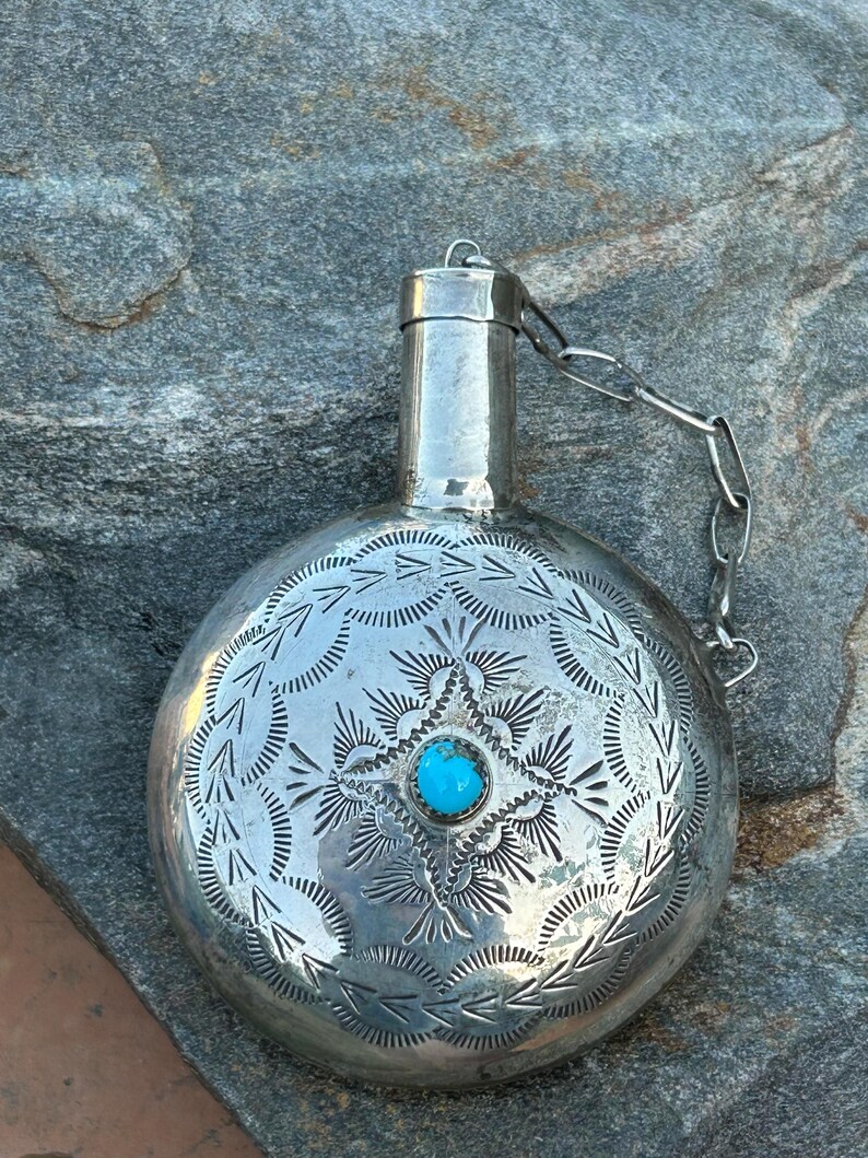 Puede incluir: Una petaca de plata con intrincados patrones grabados y una piedra turquesa central. La petaca tiene un cuerpo redondo, un cuello peque&ntilde;o y una cadena sujeta a la tapa. El dise&ntilde;o presenta motivos florales y geom&eacute;tricos detallados.