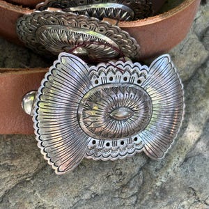 May include: Close-up of a brown leather belt with a large, ornate silver buckle. The buckle features a central oval design with radiating lines and decorative edges. The belt is resting on a textured, gray stone surface.