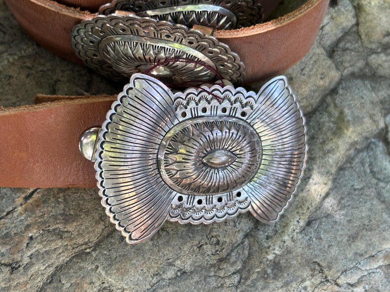 May include: Close-up of a brown leather belt with a large, ornate silver buckle. The buckle features a central oval design with radiating lines and decorative edges. The belt is resting on a textured, gray stone surface.