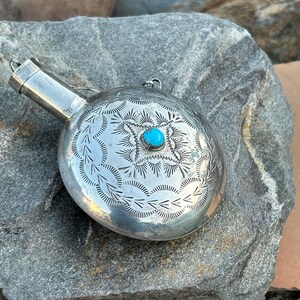Puede incluir: Una petaca plateada con intrincados dise&ntilde;os grabados y una peque&ntilde;a piedra turquesa redonda en el centro. La petaca tiene un pico y un peque&ntilde;o lazo para sujetar una cadena. El metal tiene un acabado pulido.