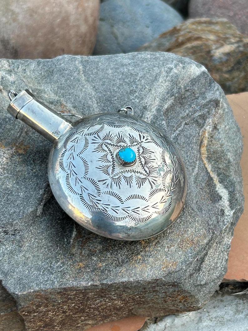 Puede incluir: Una petaca plateada con intrincados dise&ntilde;os grabados y una peque&ntilde;a piedra turquesa redonda en el centro. La petaca tiene un pico y un peque&ntilde;o lazo para sujetar una cadena. El metal tiene un acabado pulido.
