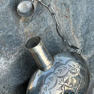 Puede incluir: Una petaca plateada con un dise&ntilde;o grabado y una peque&ntilde;a piedra turquesa. La petaca tiene una cadena unida a la tapa. La petaca met&aacute;lica tiene forma redondeada.