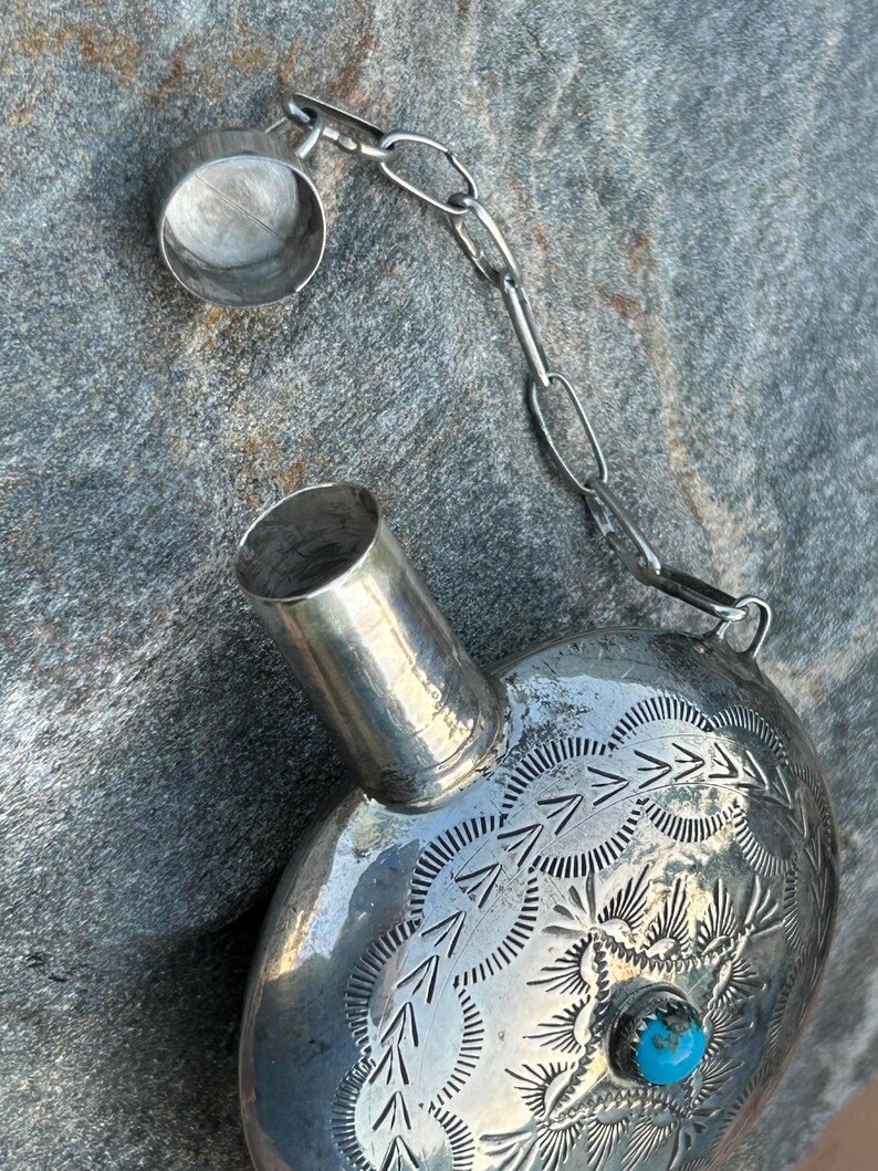 Puede incluir: Una petaca plateada con un dise&ntilde;o grabado y una peque&ntilde;a piedra turquesa. La petaca tiene una cadena unida a la tapa. La petaca met&aacute;lica tiene forma redondeada.