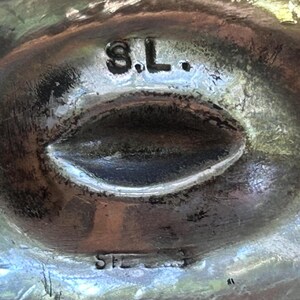 May include: Close-up of a silver-toned metal object with an oval-shaped indentation. The letters "S.L." are stamped at the top, and "STERLING" is stamped at the bottom. The object has a textured surface.