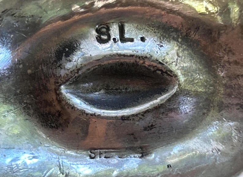 May include: Close-up of a silver-toned metal object with an oval-shaped indentation. The letters "S.L." are stamped at the top, and "STERLING" is stamped at the bottom. The object has a textured surface.