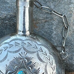 Puede incluir: Una petaca de plata con intrincados patrones grabados y una incrustaci&oacute;n de piedra turquesa. La petaca tiene un cuello cil&iacute;ndrico y est&aacute; unida a una cadena. La superficie est&aacute; adornada con dise&ntilde;os geom&eacute;tricos detallados, creando una est&eacute;tica vintage.