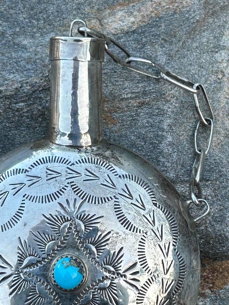 Puede incluir: Una petaca de plata con intrincados patrones grabados y una incrustaci&oacute;n de piedra turquesa. La petaca tiene un cuello cil&iacute;ndrico y est&aacute; unida a una cadena. La superficie est&aacute; adornada con dise&ntilde;os geom&eacute;tricos detallados, creando una est&eacute;tica vintage.