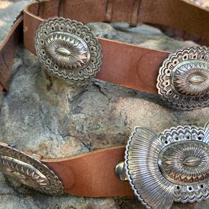 May include: A brown leather belt with multiple silver-colored decorative buckles. The buckles have an oval shape with intricate floral and sunburst designs. The belt is resting on a textured stone surface.