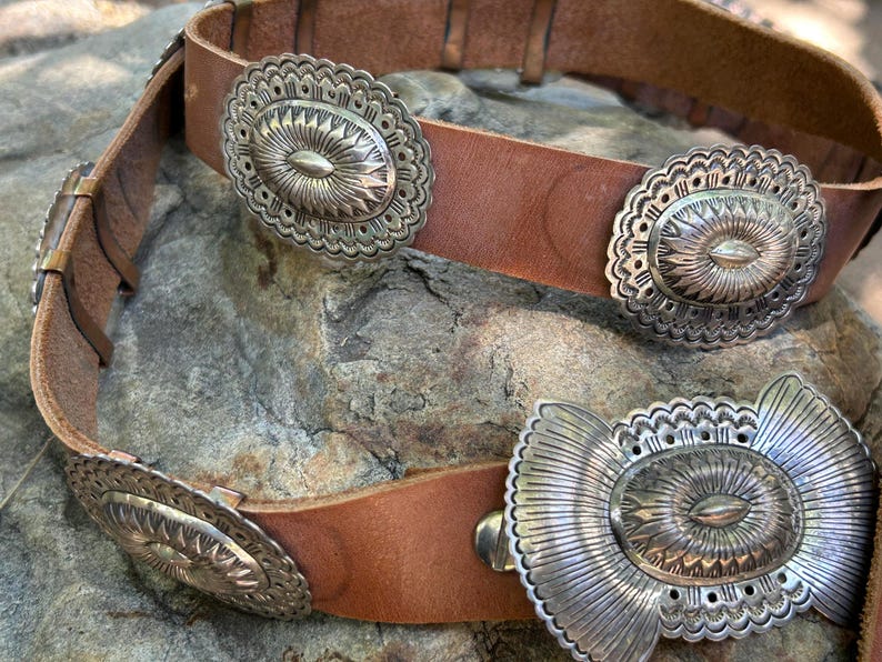 May include: A brown leather belt with multiple silver-colored decorative buckles. The buckles have an oval shape with intricate floral and sunburst designs. The belt is resting on a textured stone surface.