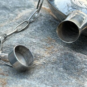 Puede incluir: Primer plano de una petaca de metal plateado con un tap&oacute;n a juego sujeto por una cadena. La petaca tiene un dise&ntilde;o decorativo en la parte superior y un pico. El tap&oacute;n est&aacute; abierto, revelando el interior de la petaca.