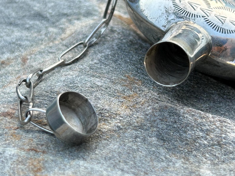 Puede incluir: Primer plano de una petaca de metal plateado con un tap&oacute;n a juego sujeto por una cadena. La petaca tiene un dise&ntilde;o decorativo en la parte superior y un pico. El tap&oacute;n est&aacute; abierto, revelando el interior de la petaca.