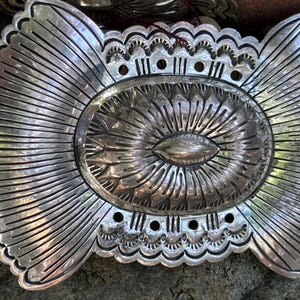 May include: A large, ornate silver belt buckle with a detailed, sunburst design. The oval center features a textured pattern, surrounded by radiating lines and scalloped edges. The buckle is likely made of metal and is designed to be attached to a belt.