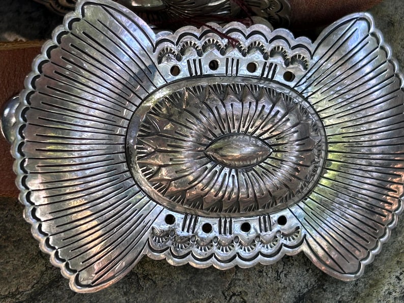 May include: A large, ornate silver belt buckle with a detailed, sunburst design. The oval center features a textured pattern, surrounded by radiating lines and scalloped edges. The buckle is likely made of metal and is designed to be attached to a belt.
