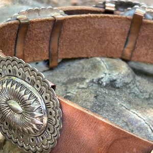 May include: Close-up of a brown leather belt with silver-colored decorative elements. The belt features an oval buckle with a sunburst design and scalloped edges. The belt is a western-style accessory.