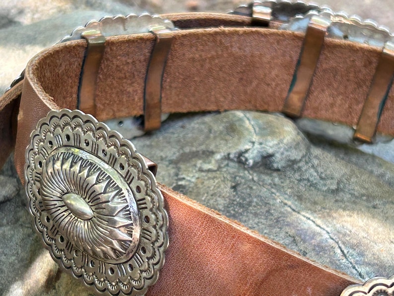 May include: Close-up of a brown leather belt with silver-colored decorative elements. The belt features an oval buckle with a sunburst design and scalloped edges. The belt is a western-style accessory.