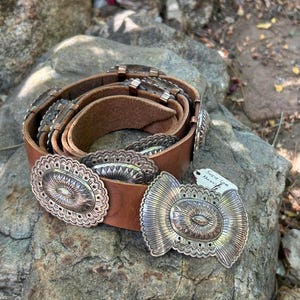 May include: A brown leather belt with ornate silver-colored metal embellishments. The belt features oval and butterfly-shaped buckles with intricate designs. The belt is displayed on a textured rock surface.
